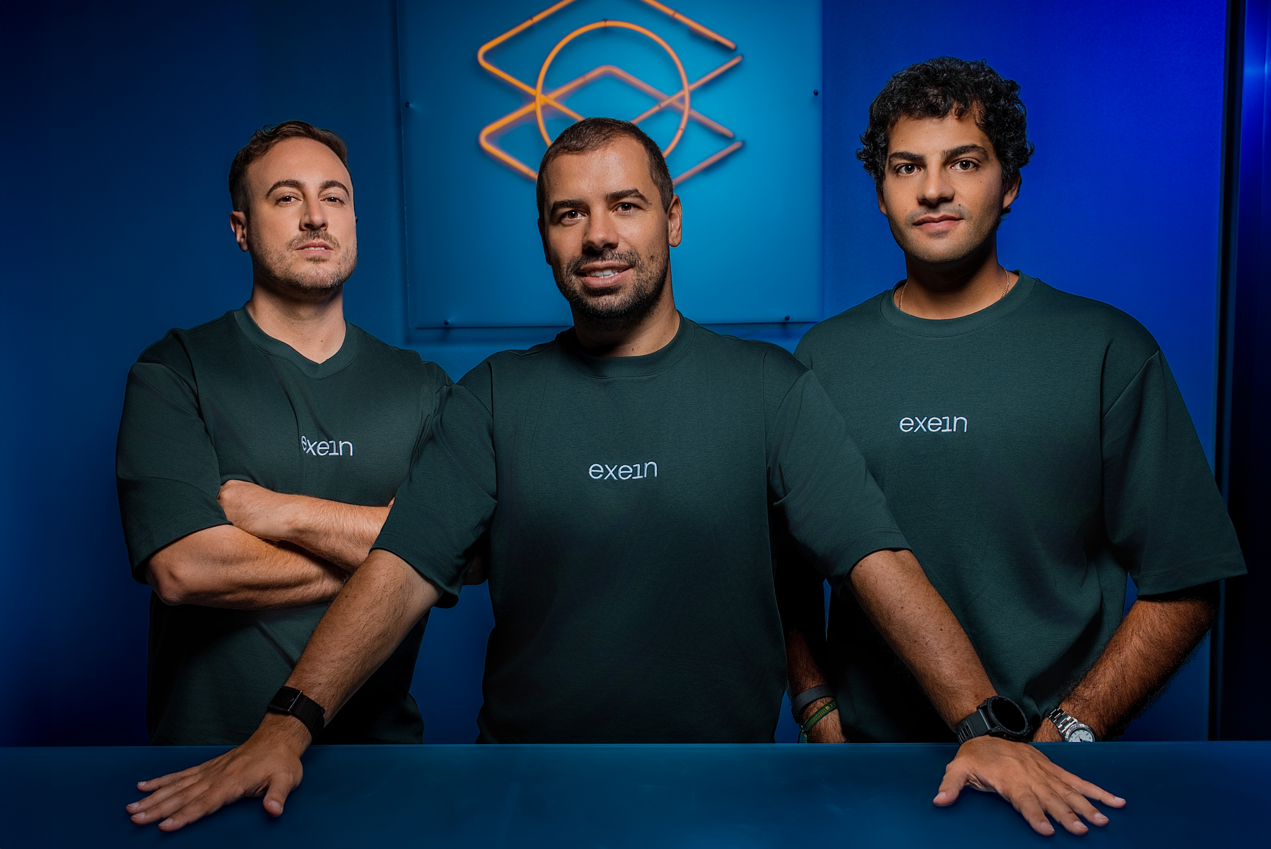 Three men in matching dark Exein shirts stand behind a blue counter against a blue backdrop.