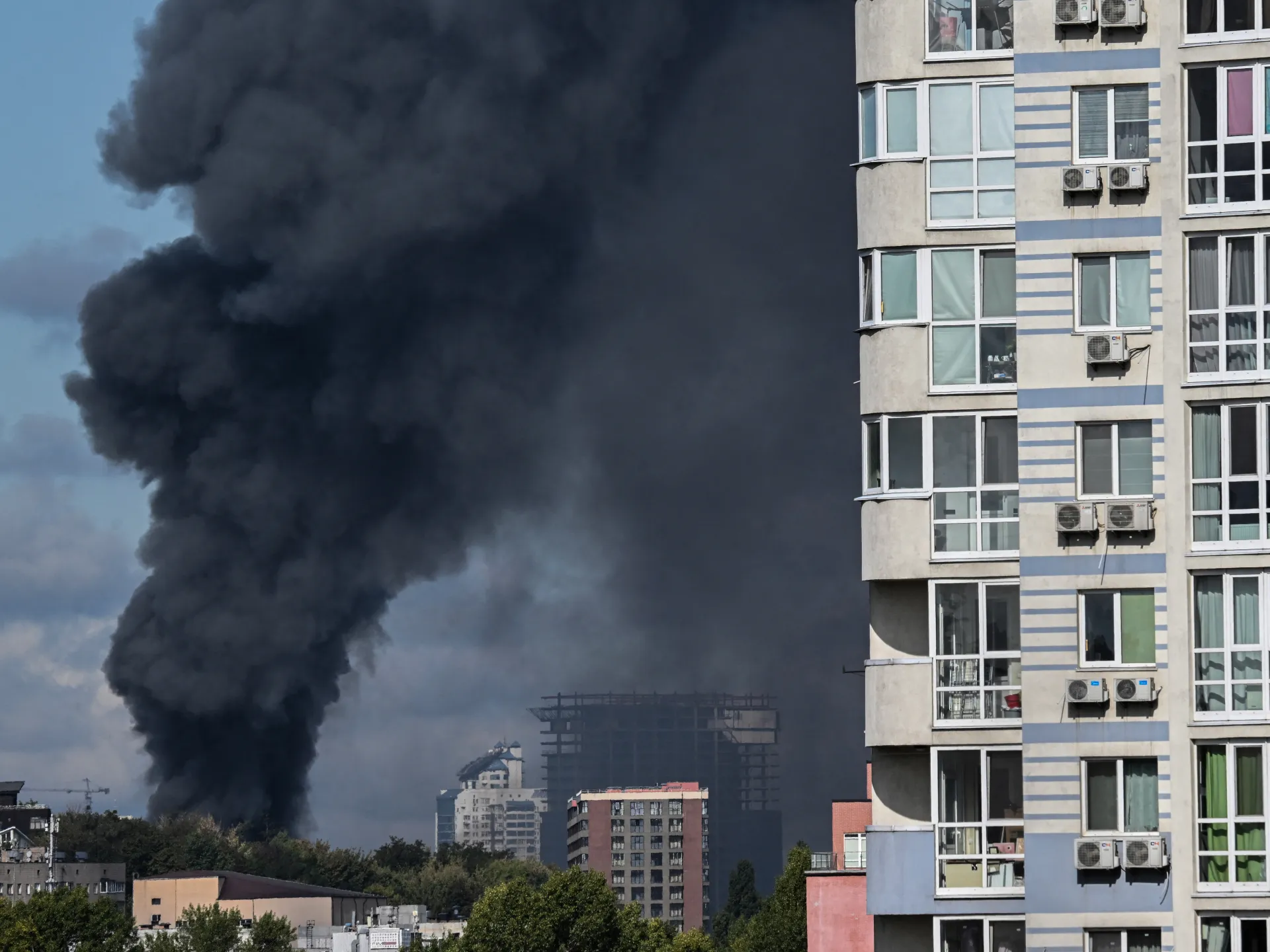 A large plume of dark smoke rises behind low buildings beside a tall apartment block.