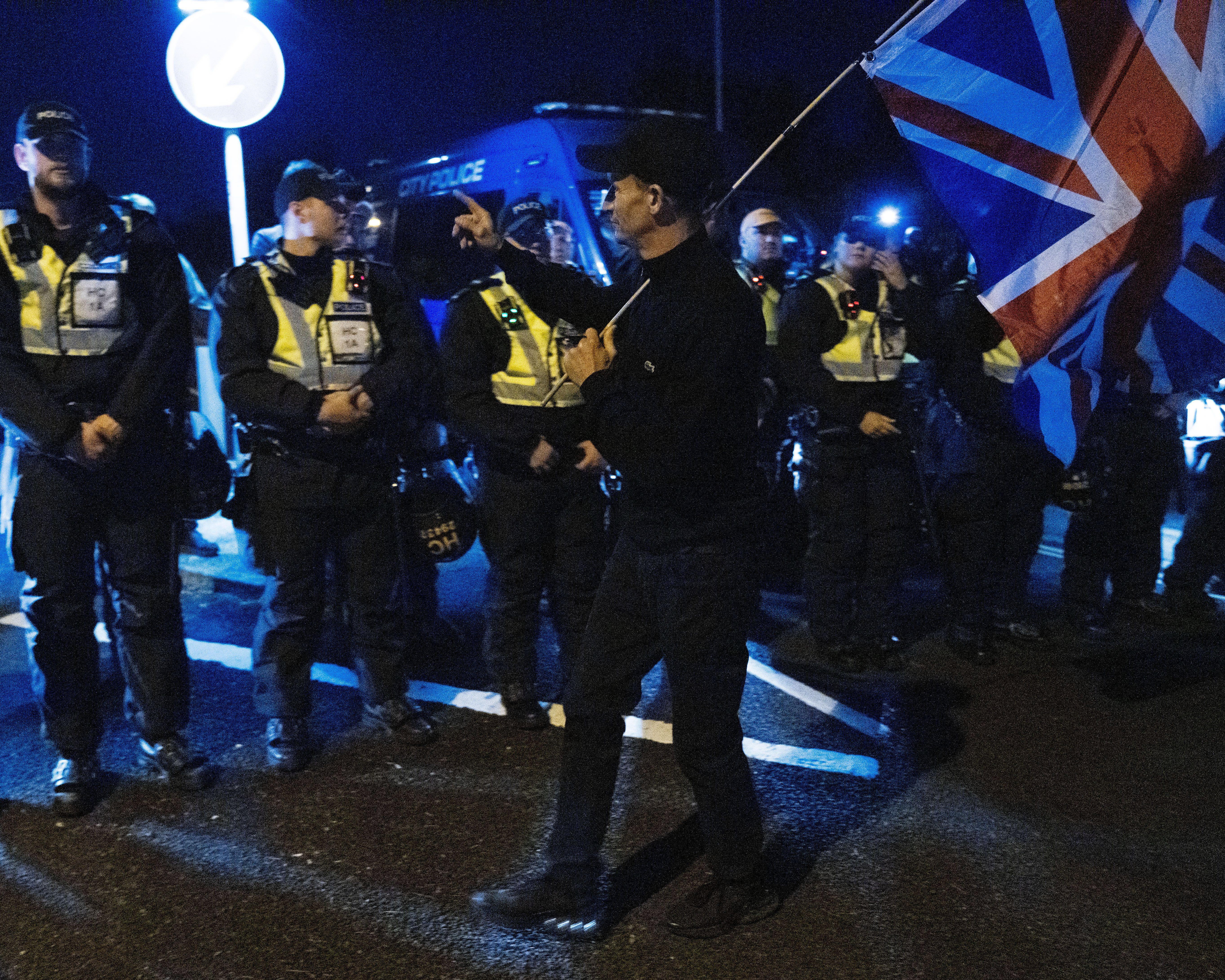 Police officers in dark uniforms with yellow panels stand beside a large Union Jack flag at night.