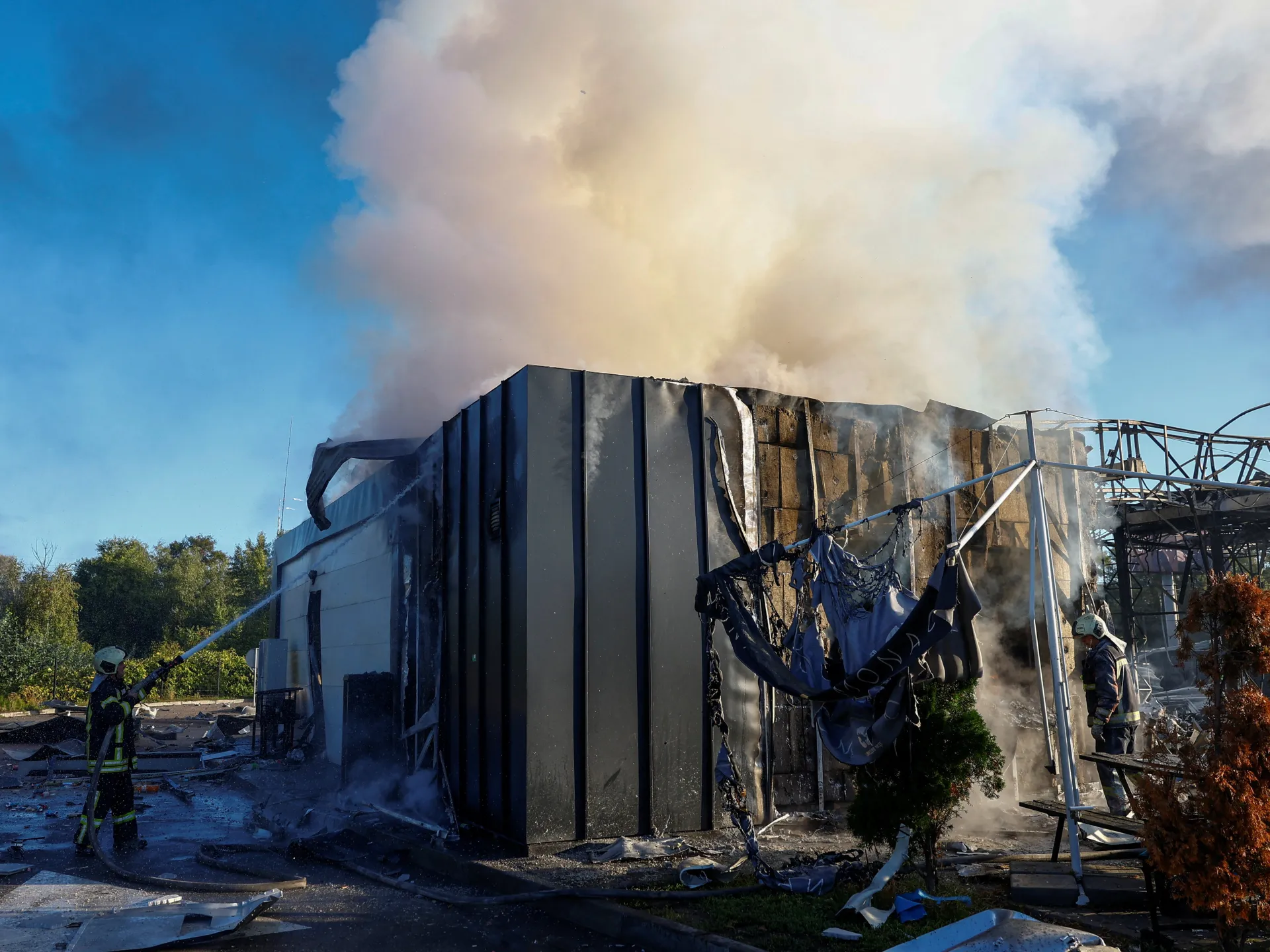 Firefighters and crane operators work beside a damaged industrial building as smoke rises into the sky.