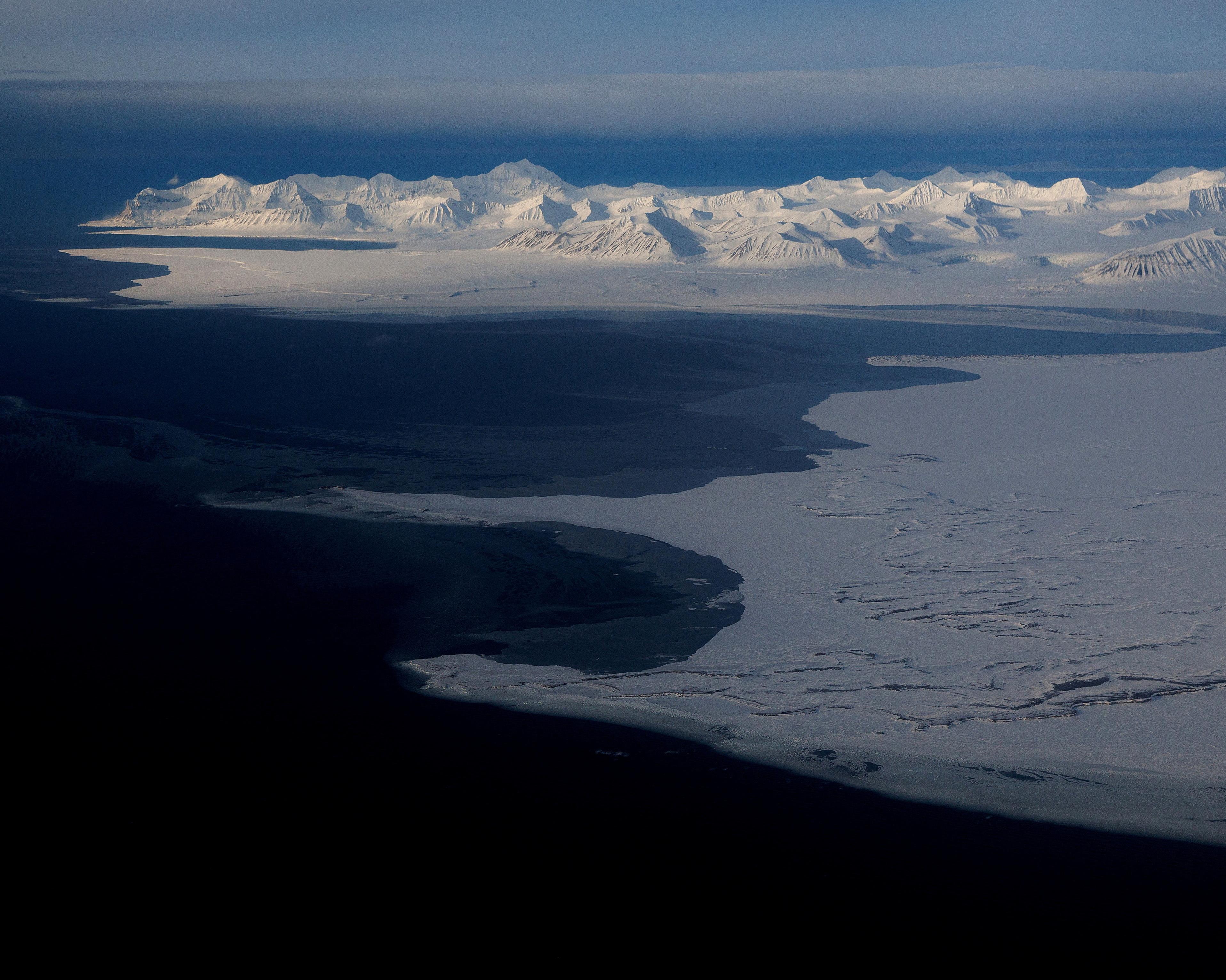 Aerial view of dark ocean water beside pale ice-covered land and distant snowy mountains.