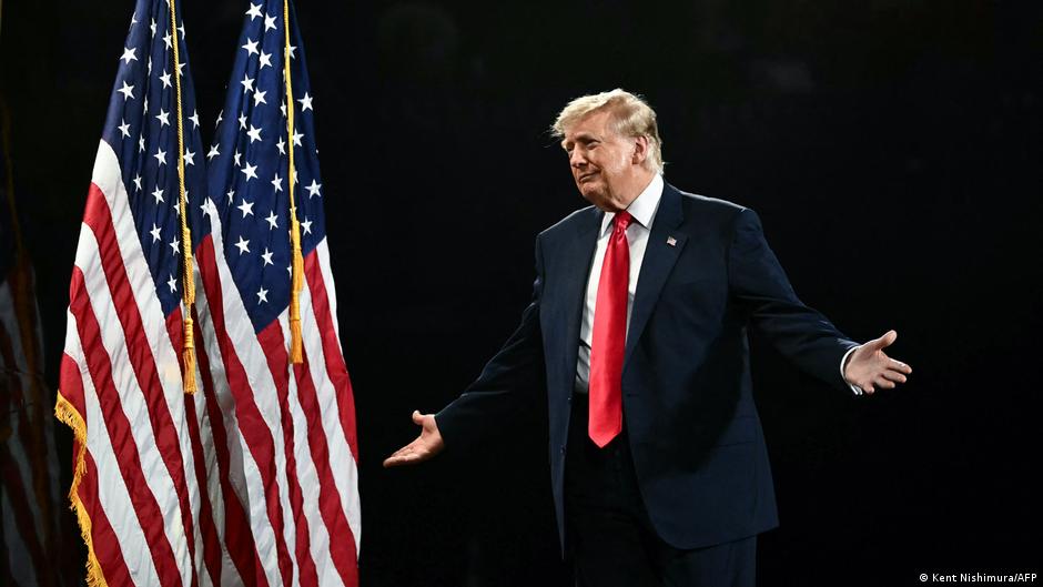 Donald Trump stands with arms outstretched beside several American flags against a dark background.