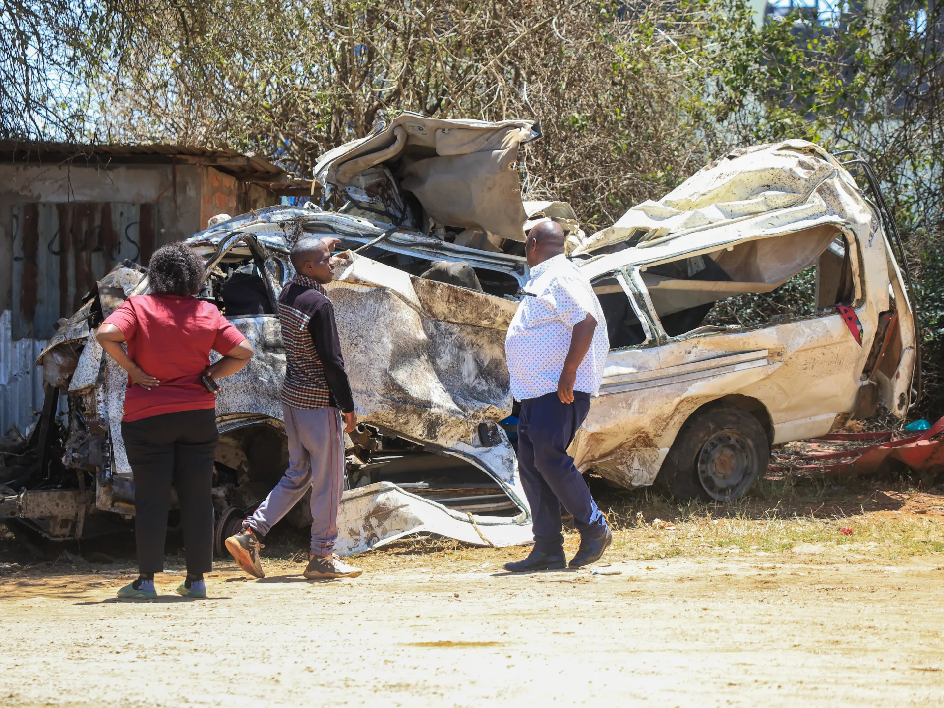 Three people stand beside a severely wrecked vehicle in a sunlit outdoor area.