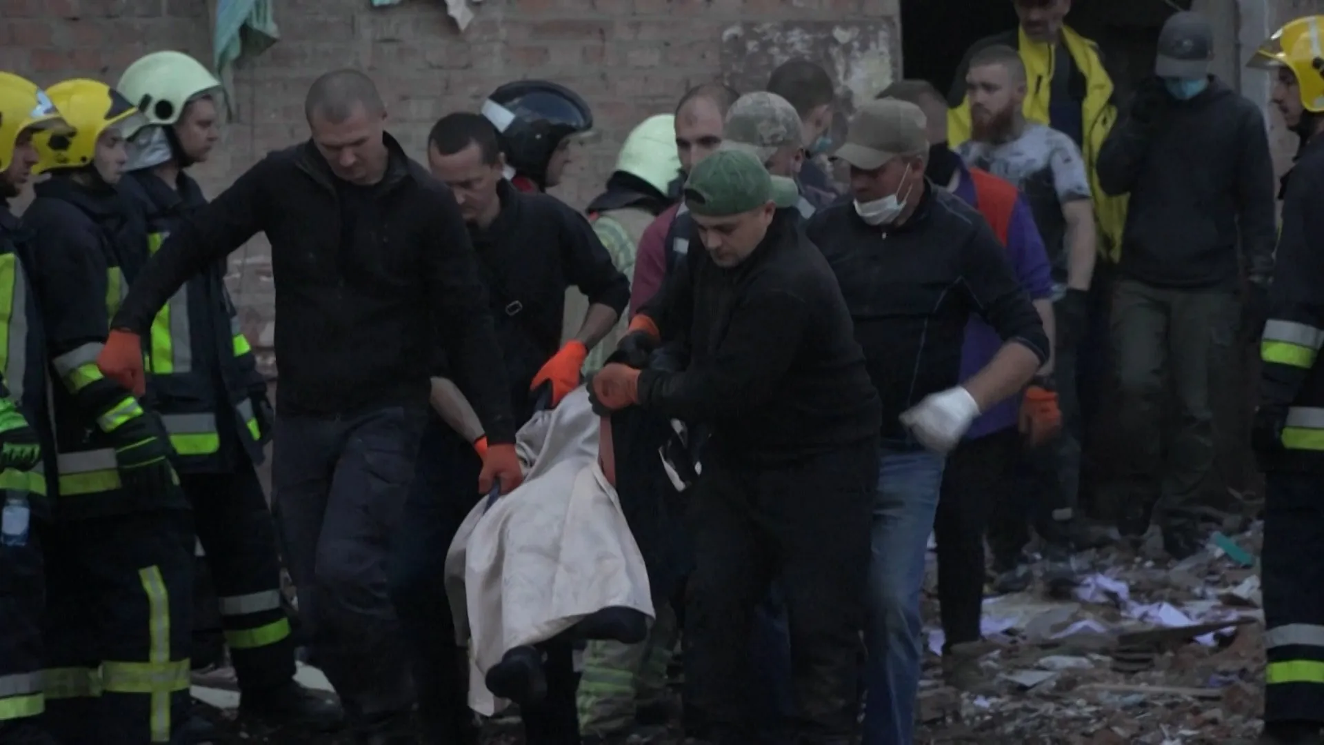Rescue workers and civilians carry a person covered by a white cloth through rubble.