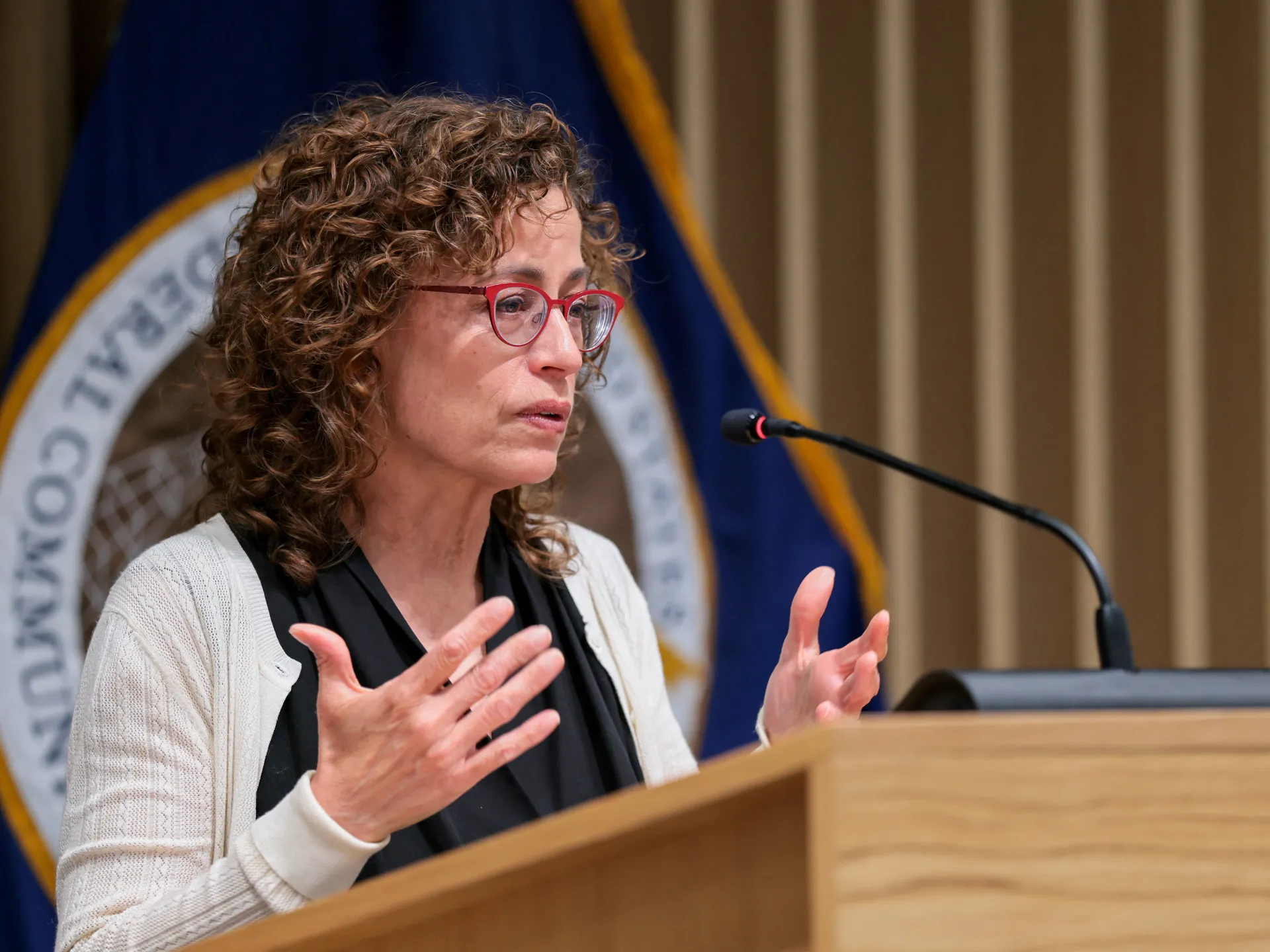 A woman in red glasses speaks at a wooden podium, gesturing beside a microphone.