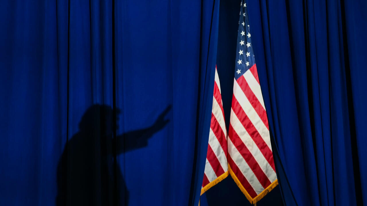 A shadowed figure gestures beside two U.S. flags against a deep blue curtain.