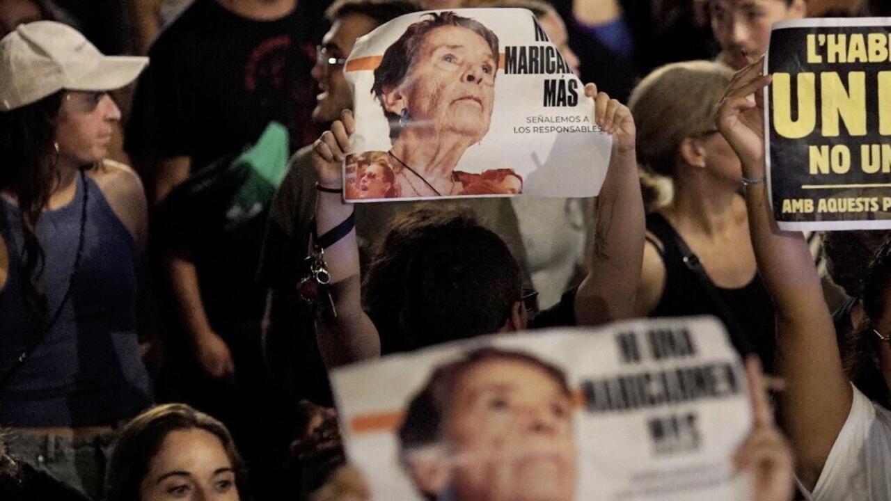 Protesters hold signs, including a portrait of an older woman, in a nighttime crowd.