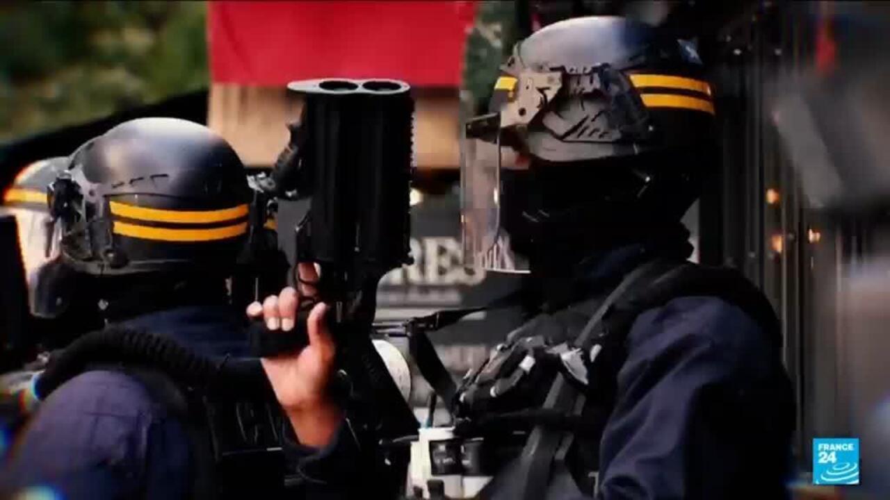 Two helmeted riot police officers stand near a vehicle, one holding a large black launcher.