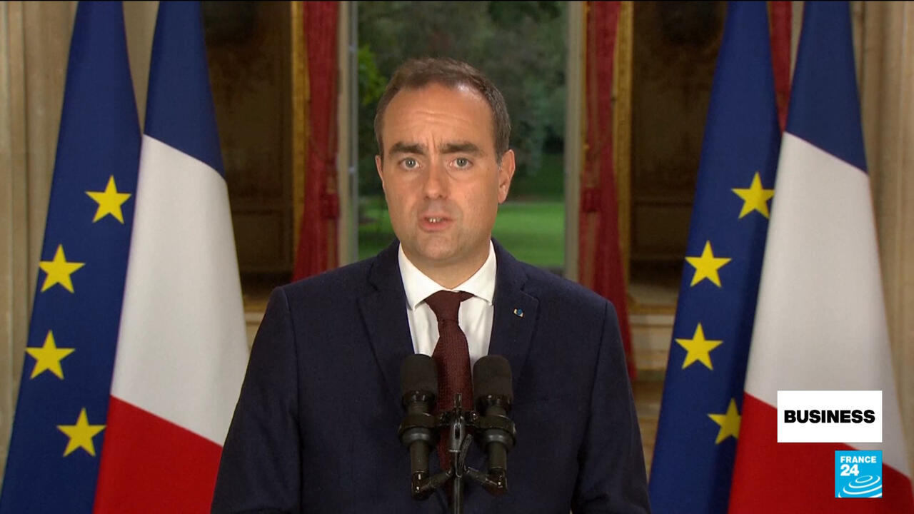 A suited man speaks at microphones between French and European Union flags.