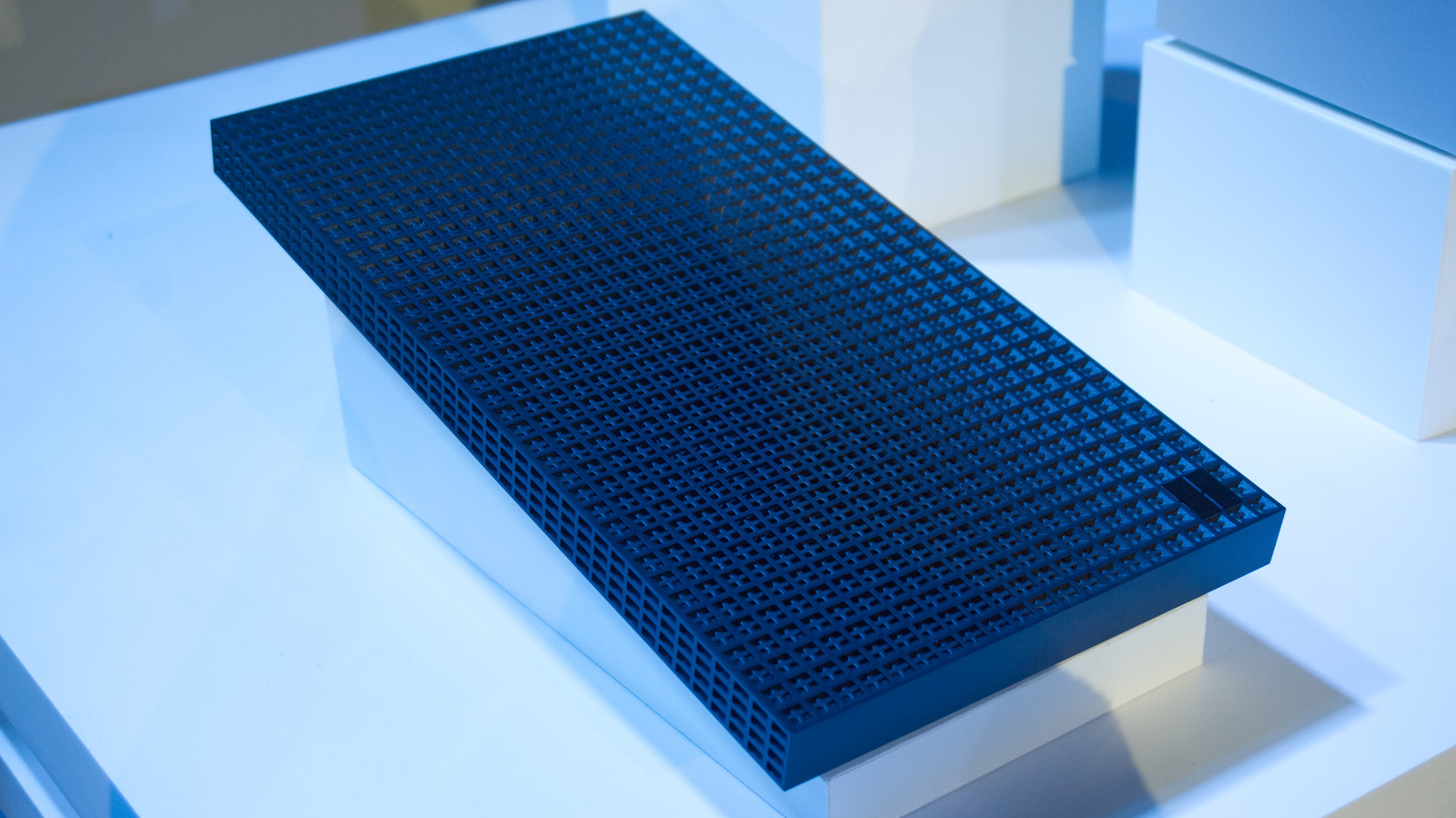 A black rectangular computer unit with a dense perforated grid sits on a white display platform.