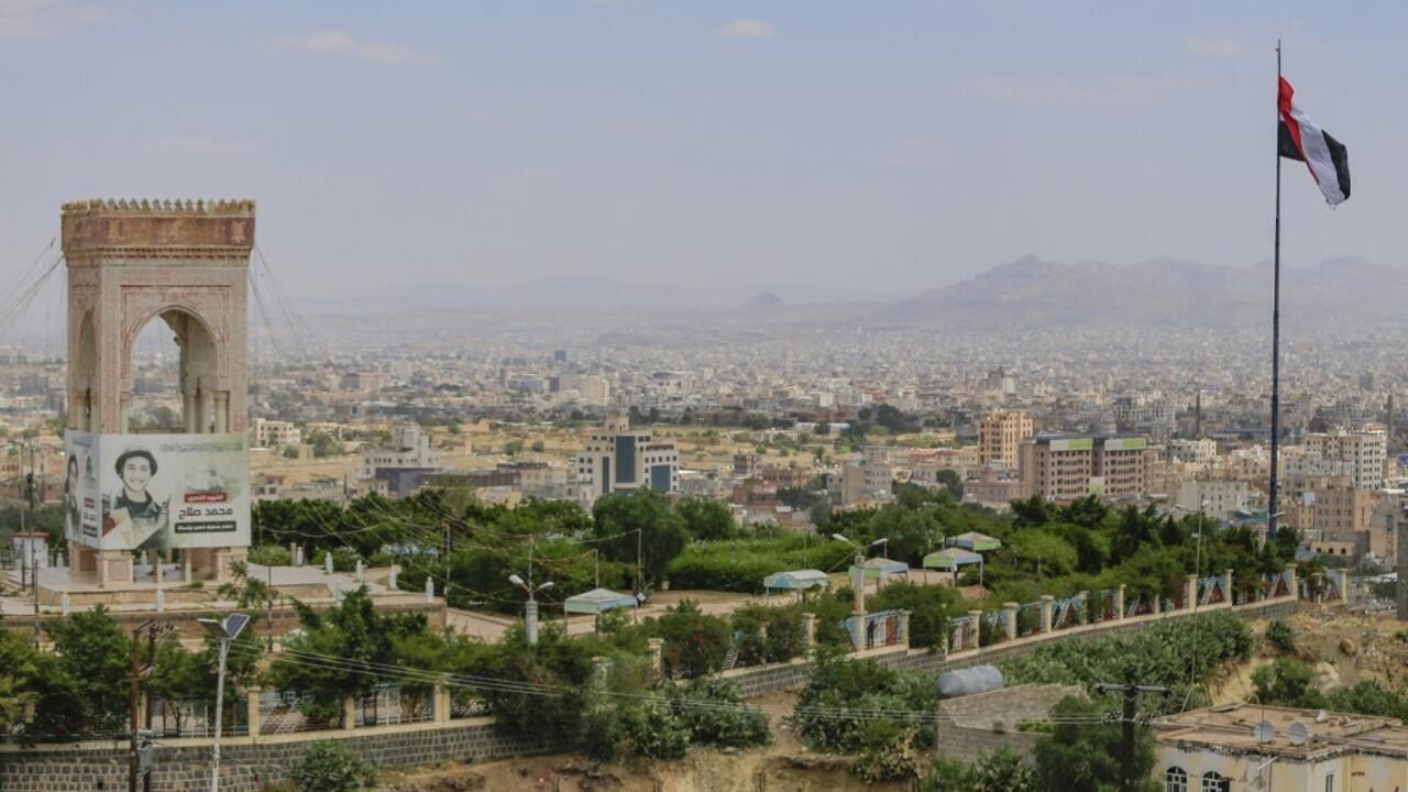 A Yemeni flag flies above a densely built city, with an ornate stone arch in the foreground.