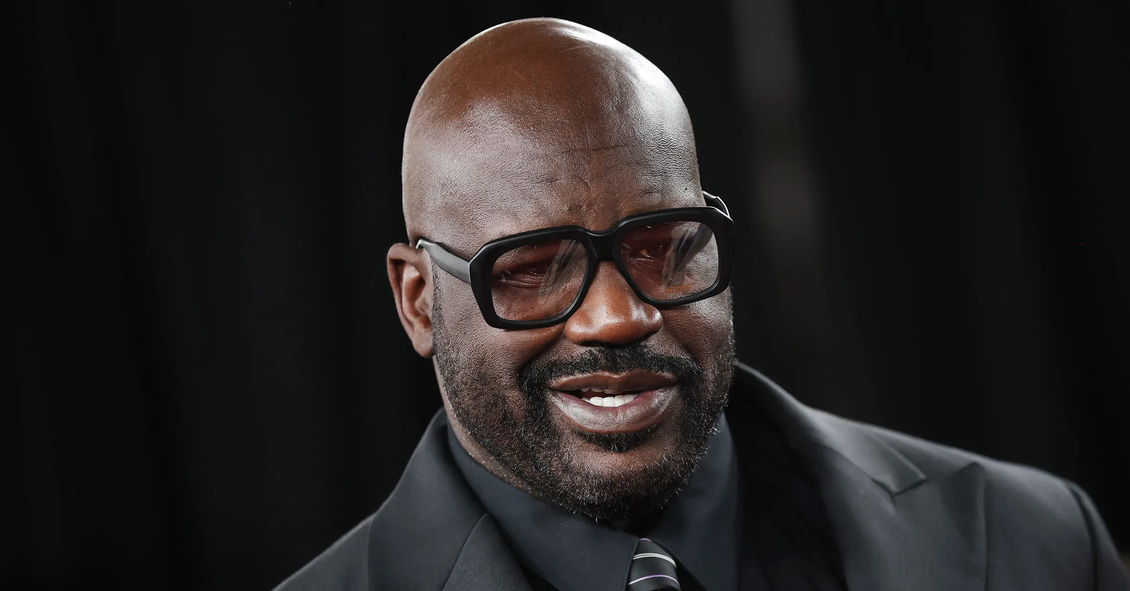 A bald man wearing oversized black glasses, a dark suit, and a striped tie against a black background.