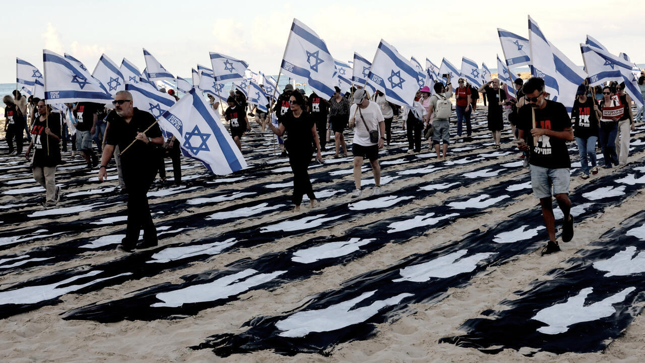 People carrying Israeli flags walk among black-and-white fabric laid across a sandy beach.