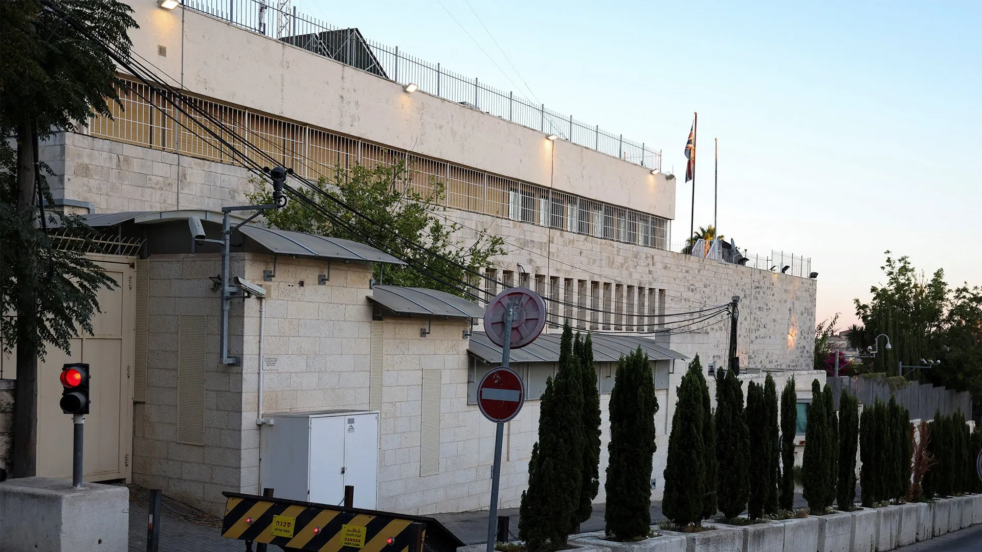 Stone building behind security barriers, with a Union Jack flag, traffic light, trees and overhead wires.