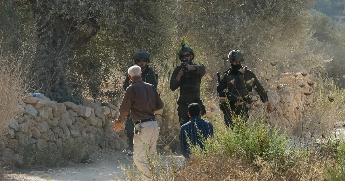 Three armed soldiers stand near two men beside a stone wall on a dry, brush-covered path.