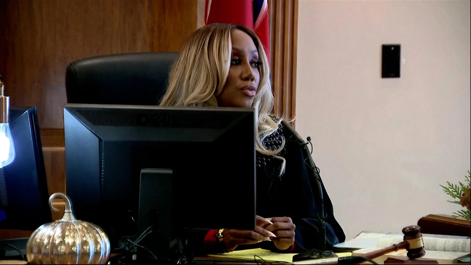 A woman sits behind a computer monitor at a desk with a gavel and documents.