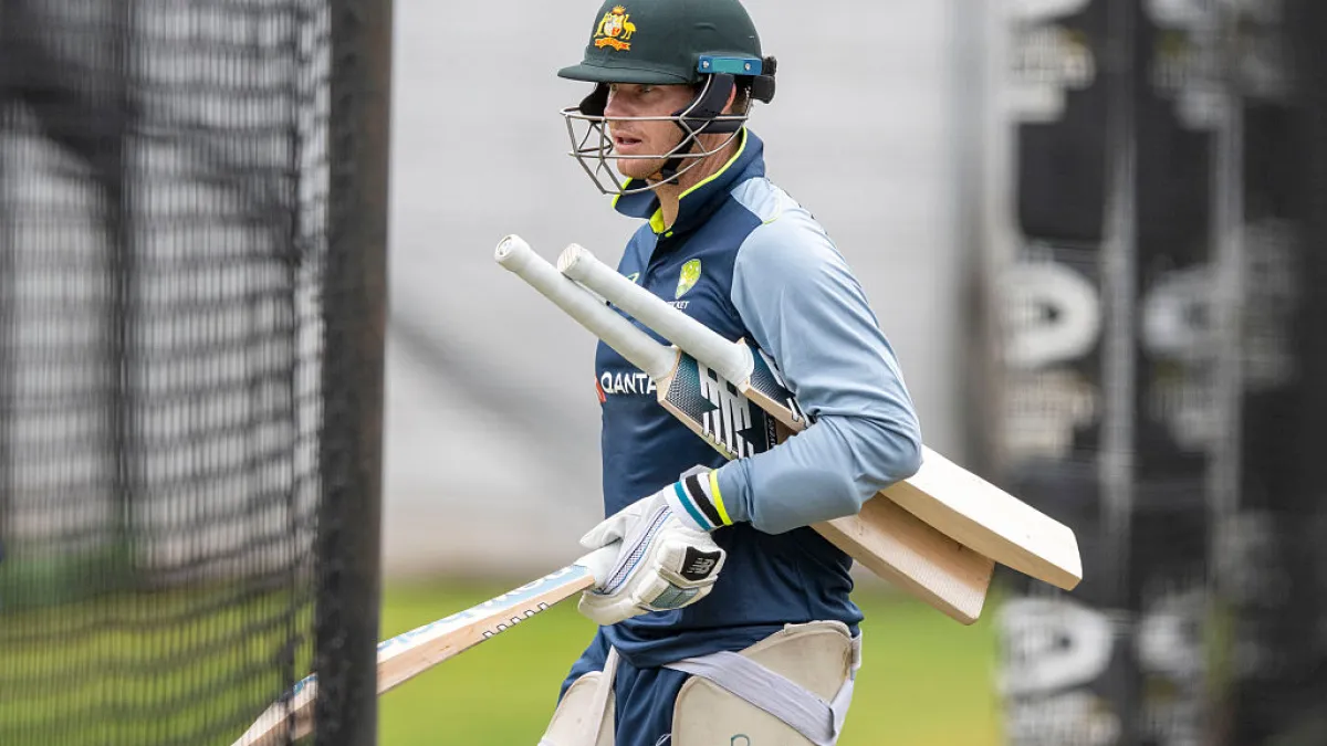 Australian cricketer in helmet and training kit carries bats while walking beside practice nets.