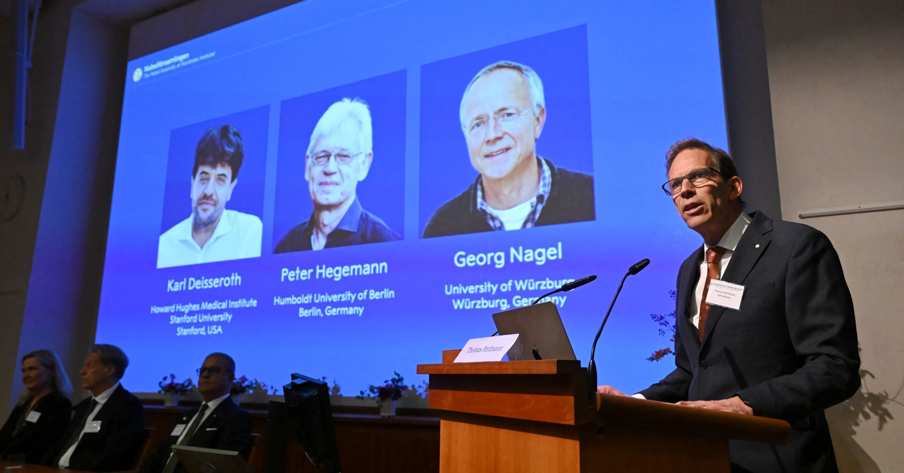 A speaker stands at a lectern beside a projected slide showing three scientists’ portraits and names.
