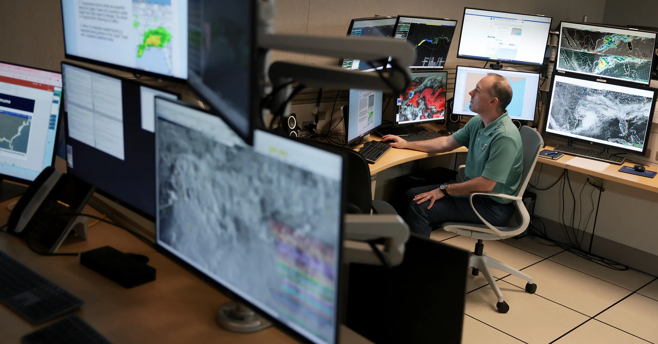A man monitors weather maps at a workstation surrounded by screens in a control room.