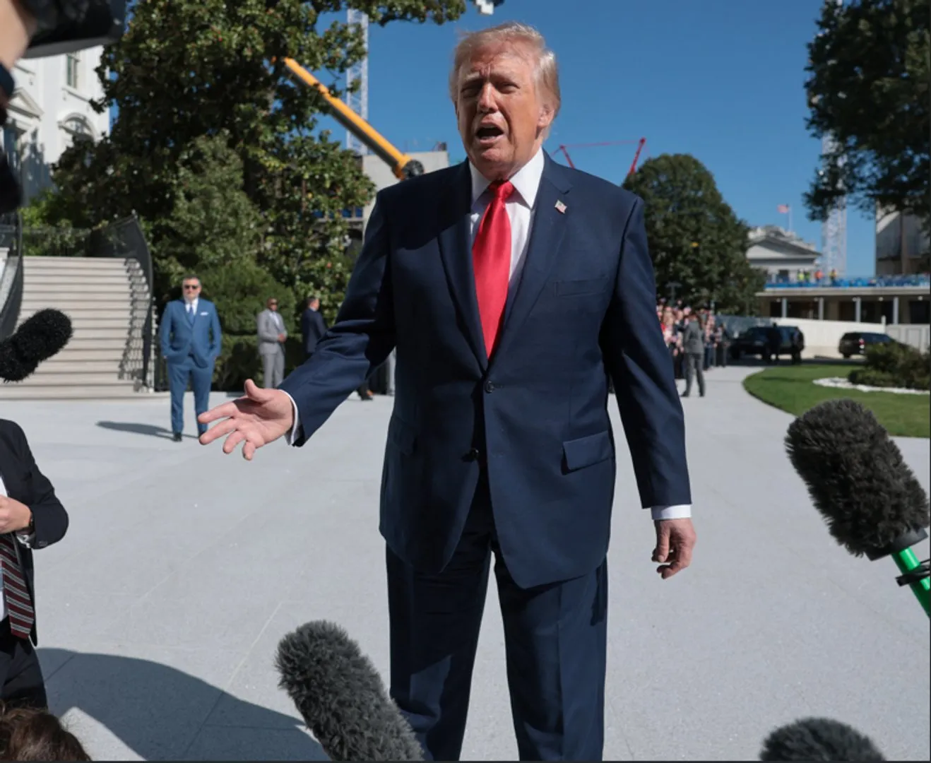 Man in a navy suit and red tie speaks outdoors, gesturing as microphones point toward him.