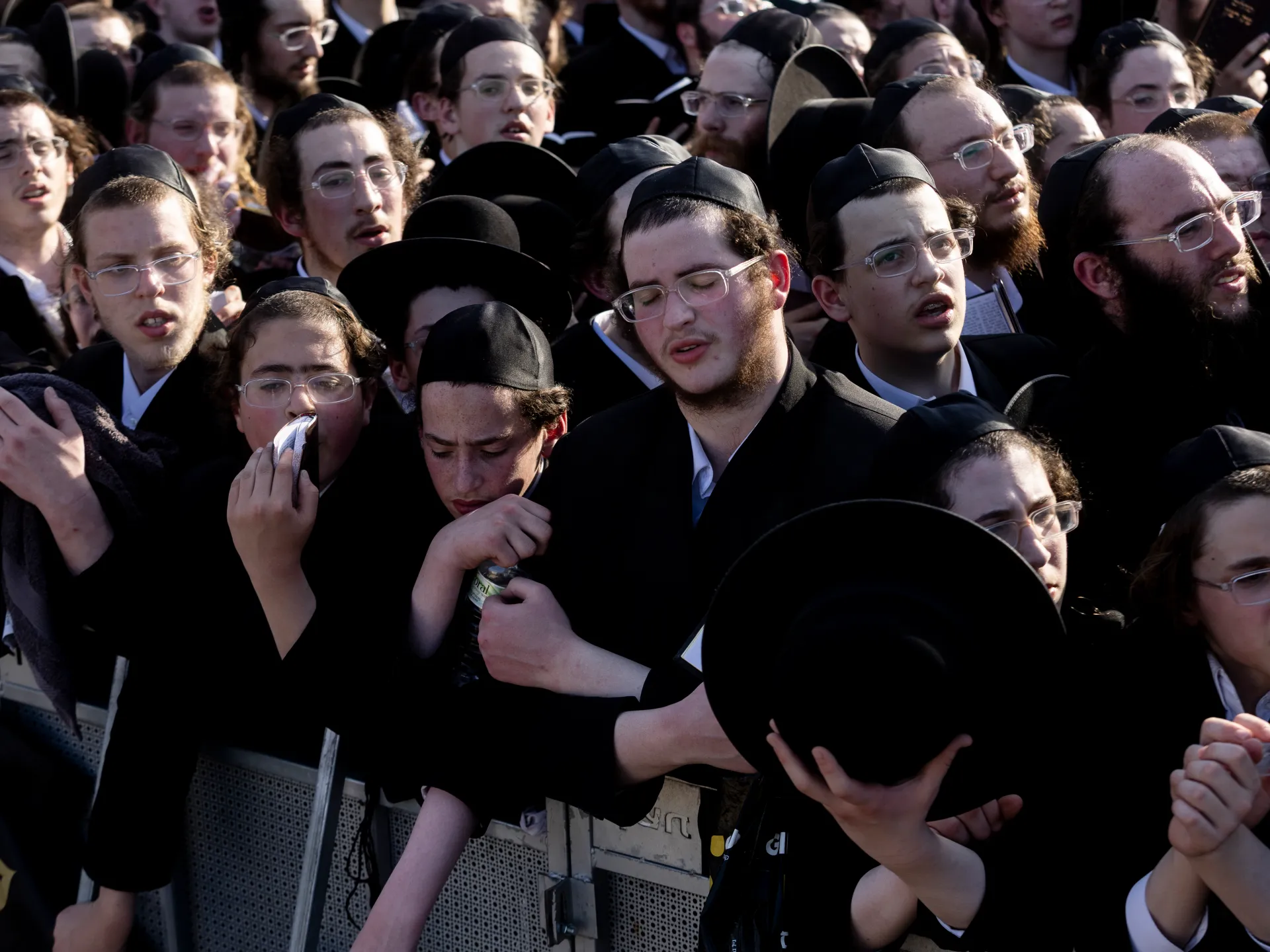 A dense crowd of ultra-Orthodox Jewish men in black clothing and hats stands behind a metal barrier.