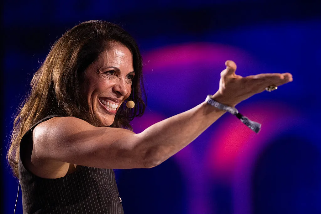 A smiling woman in a sleeveless black top gestures toward the right on a blue and magenta-lit stage.