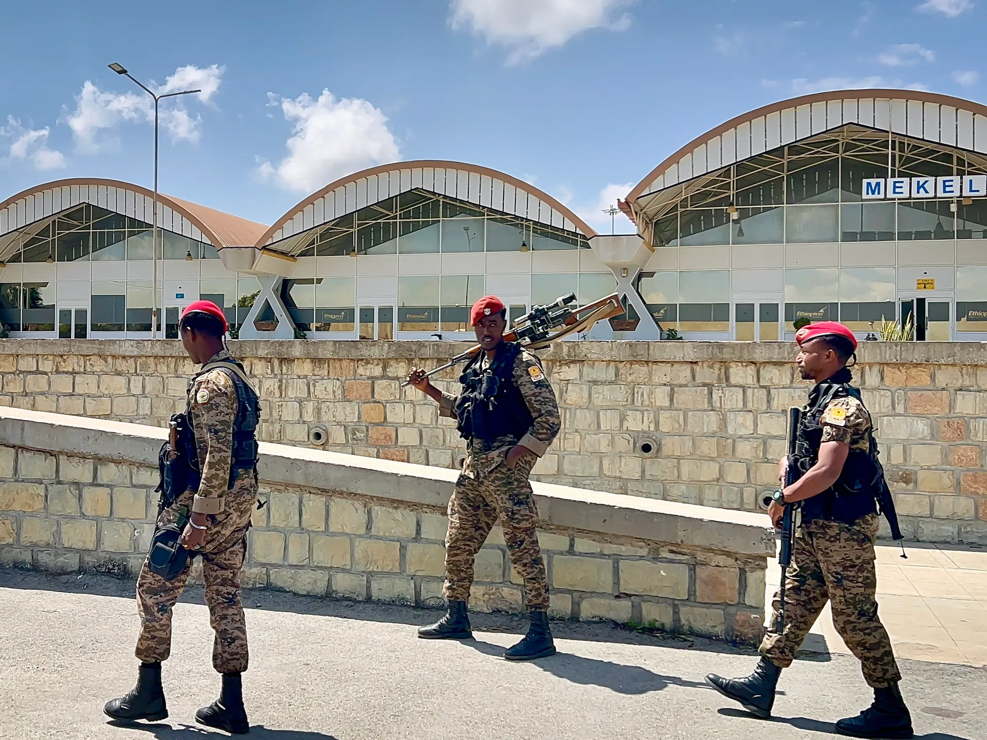 Three uniformed soldiers in red berets walk beside a stone wall and arched terminal building.