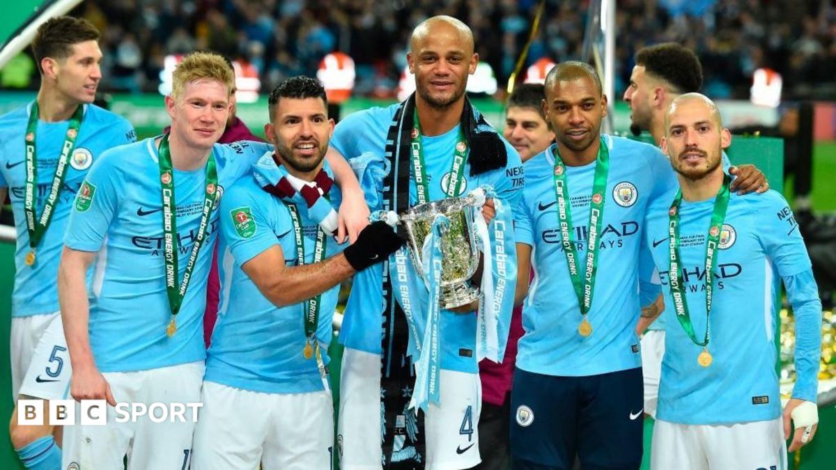 Football players in sky blue shirts pose with medals around a large silver trophy.