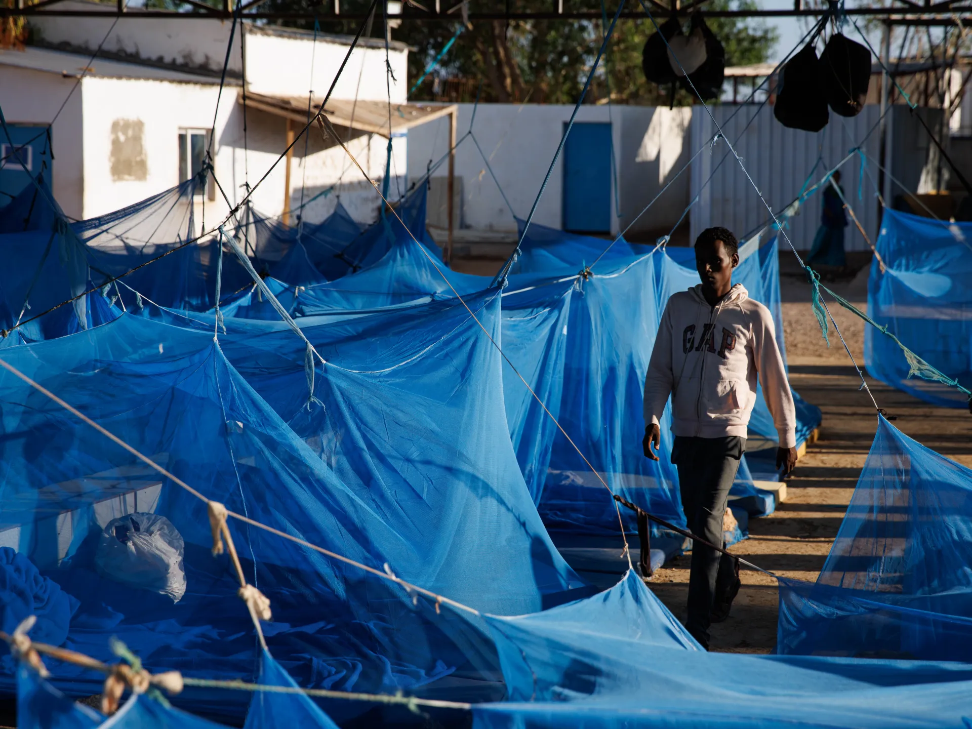A man walks among blue mosquito-net shelters in a sunlit outdoor displacement camp.