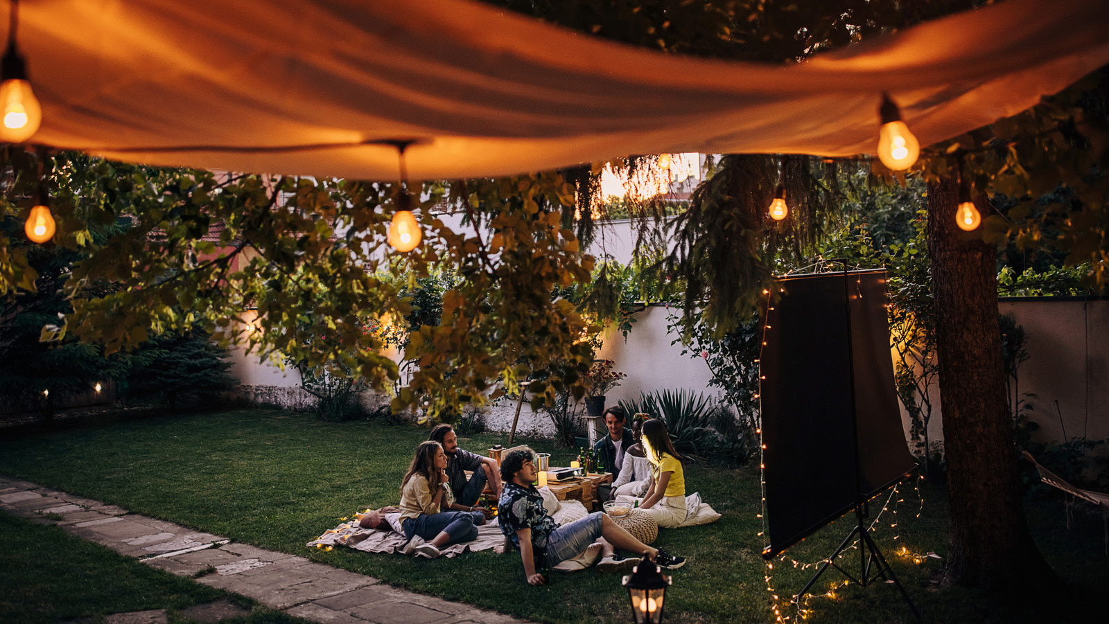 Friends gather on blankets in a warmly lit backyard beneath a canopy with string lights.