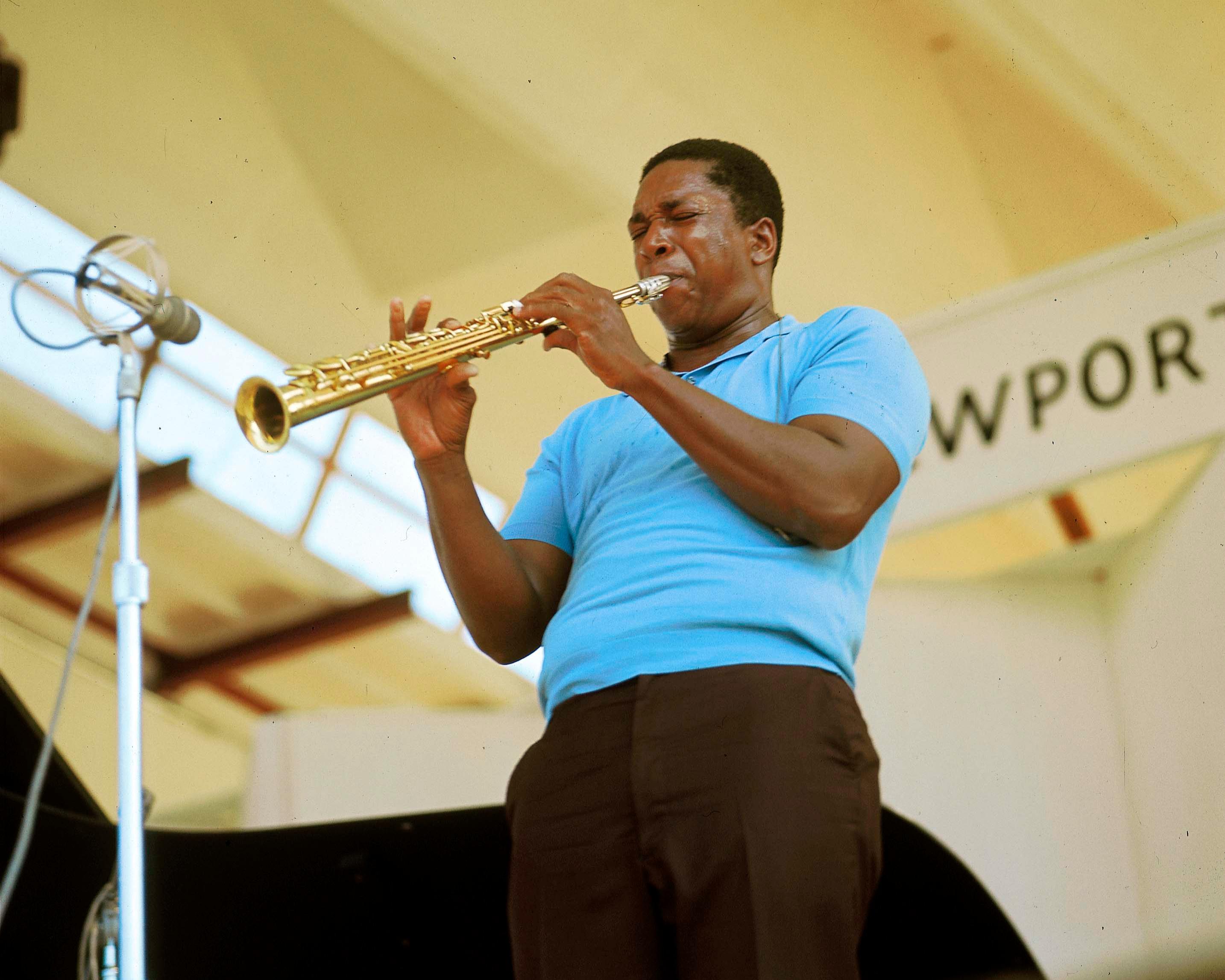 A saxophonist in a blue shirt performs onstage, playing into a microphone.