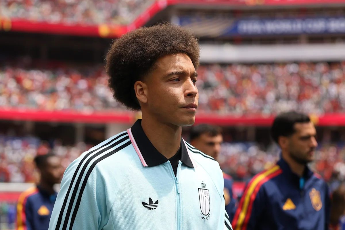 Footballer in a pale blue Adidas jacket stands on a stadium pitch before a match.