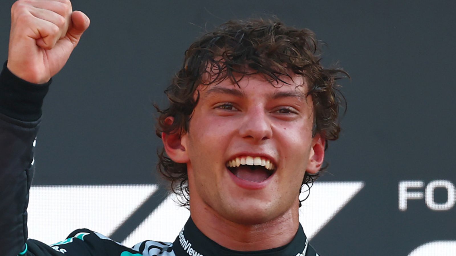 Smiling young racing driver raises a clenched fist in celebration during a podium moment.