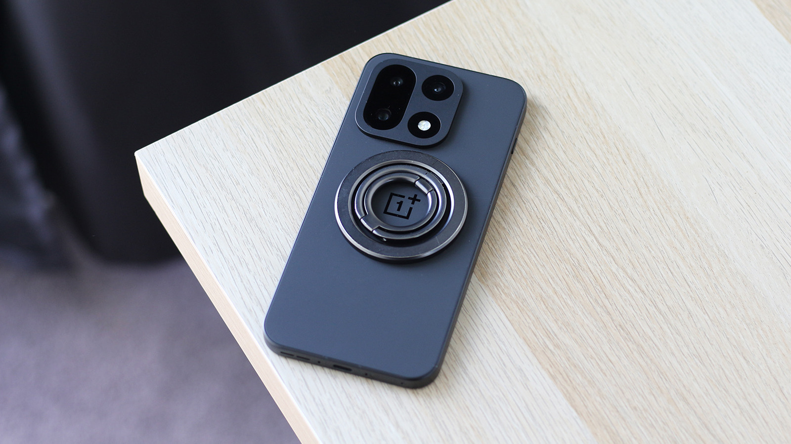 A dark smartphone with a circular magnetic ring rests diagonally on a light wooden table.