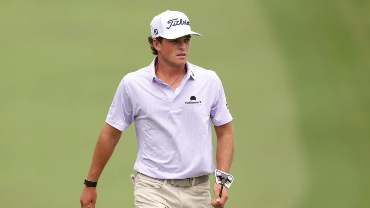 Golfer in a white cap and polo walks across the course holding a putter