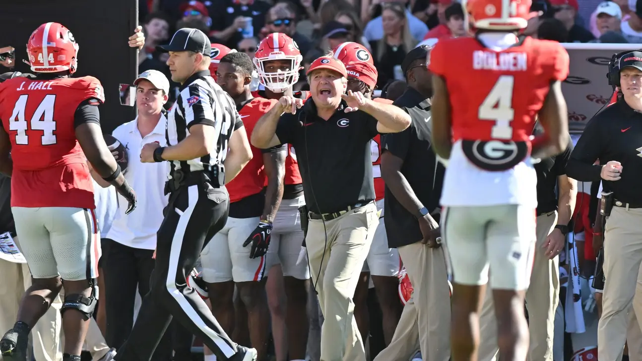 Georgia players and coaches gather on the sideline during a college football game, with a referee nearby.