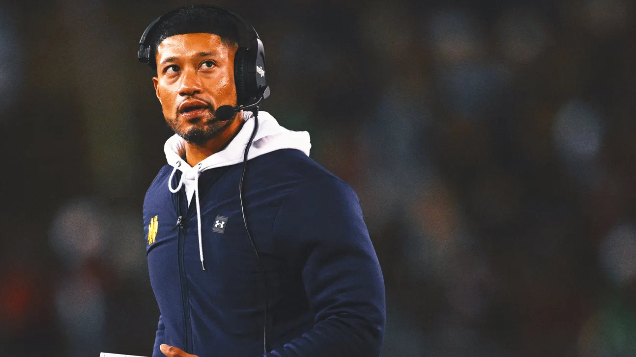 Football coach wearing a headset and navy hoodie stands on the sideline during a game.