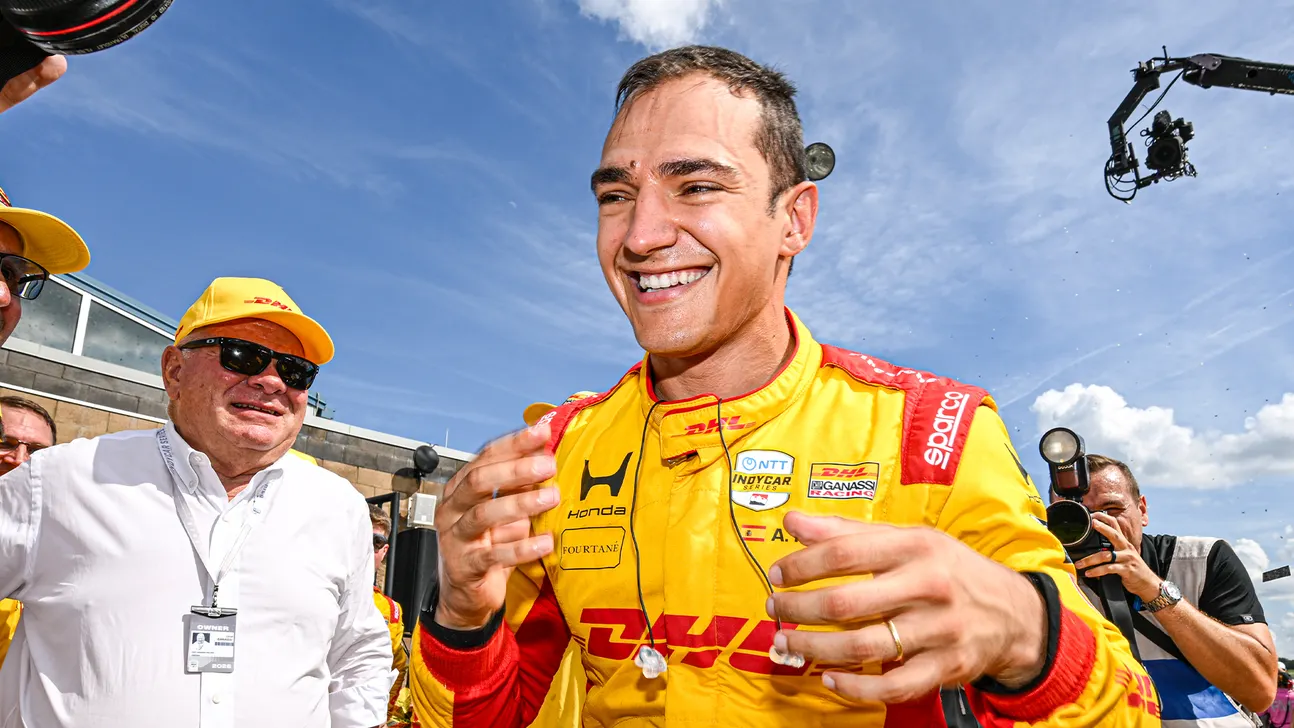 Smiling IndyCar driver in a yellow DHL racing suit gestures while photographers surround him outdoors.