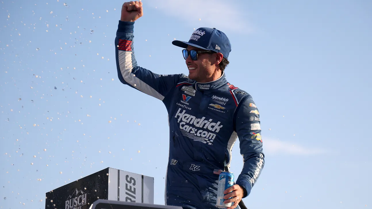 NASCAR driver in racing suit raises a fist amid confetti while holding a beverage on a podium
