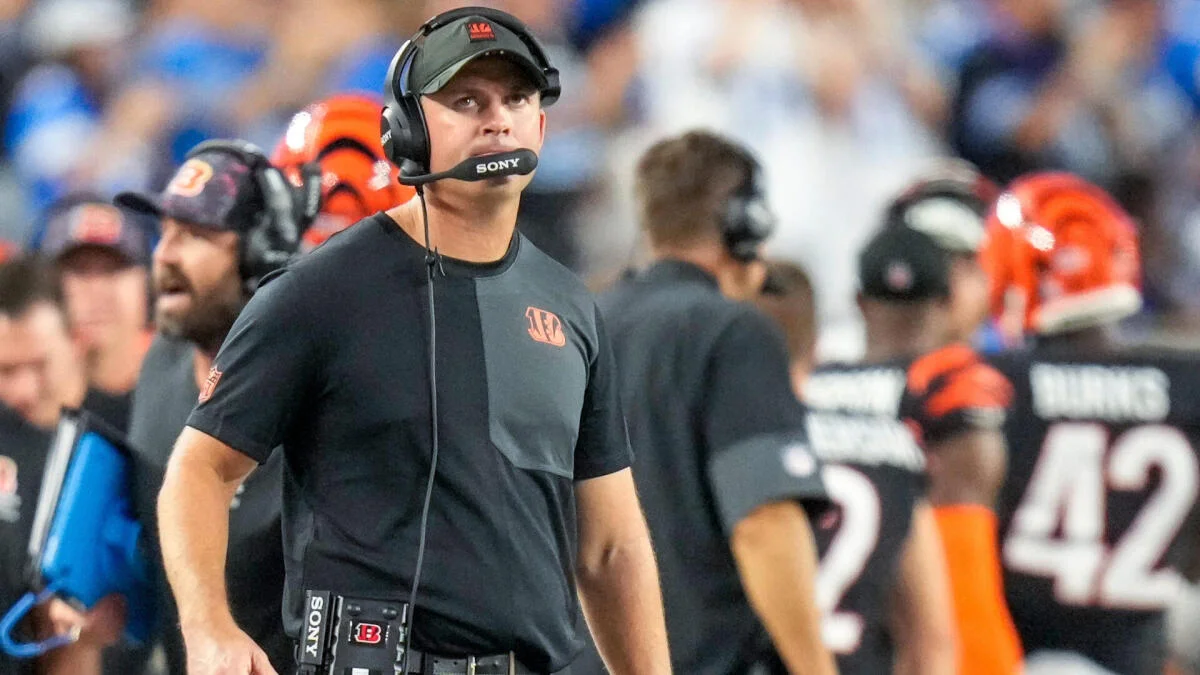 Football coach wearing headset and black shirt stands on sideline during a game.
