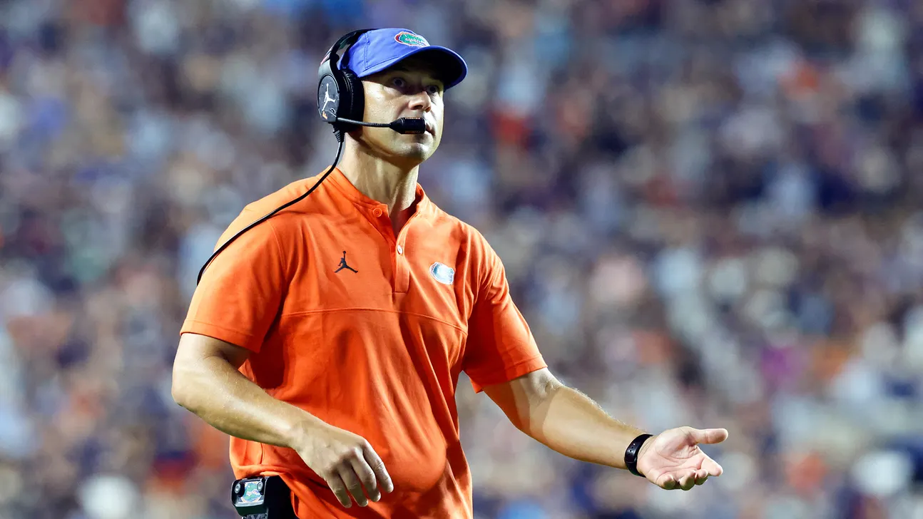 Football coach wearing an orange polo, blue cap, and headset gestures toward the field before a blurred crowd.