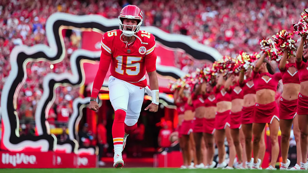 Football quarterback in red uniform running onto field as cheerleaders perform behind him