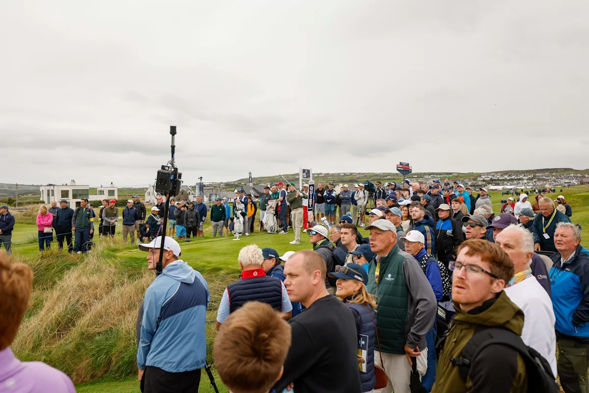 Large crowd gathers beside a golf course under an overcast sky, with a camera crane among spectators.