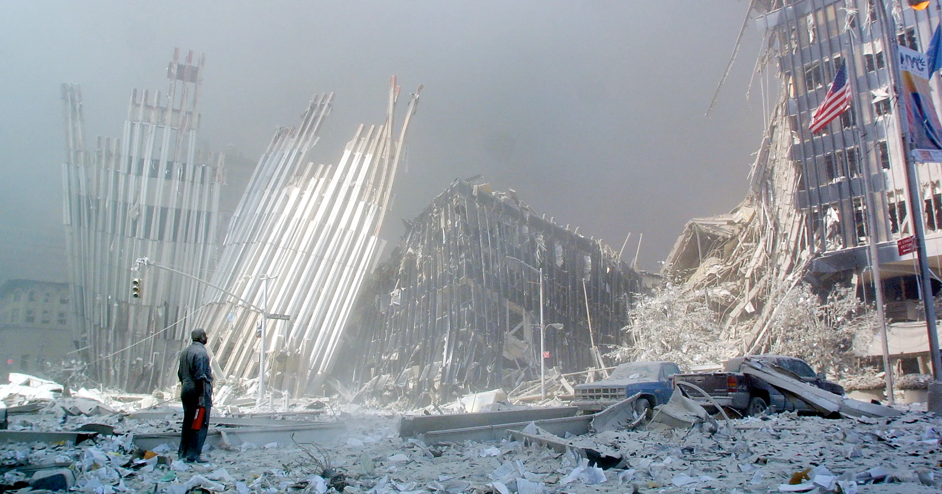 Person surveys rubble and severely damaged high-rise buildings beneath a dusty sky.