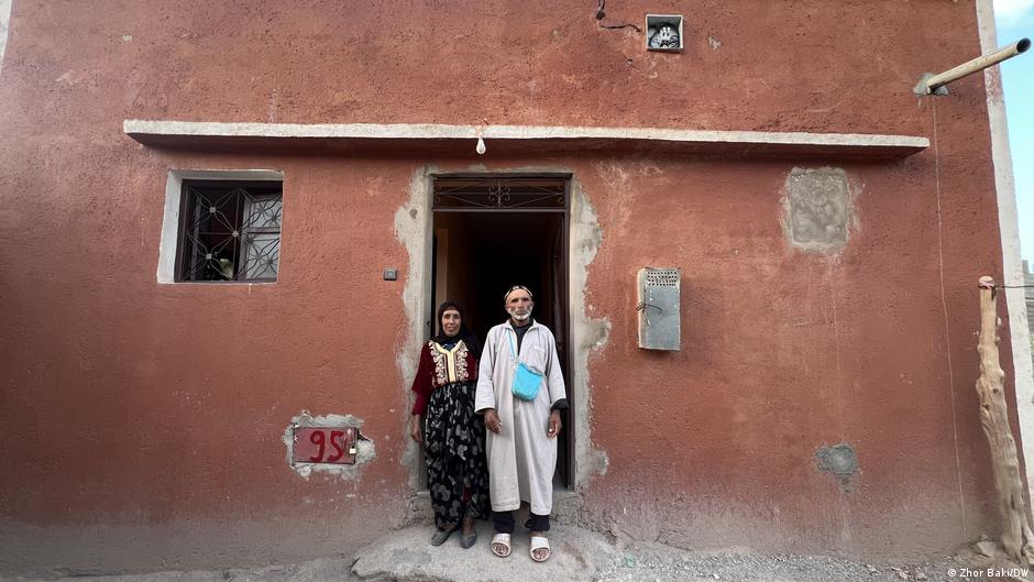 Two people stand in the doorway of a weathered reddish-brown house with barred windows.