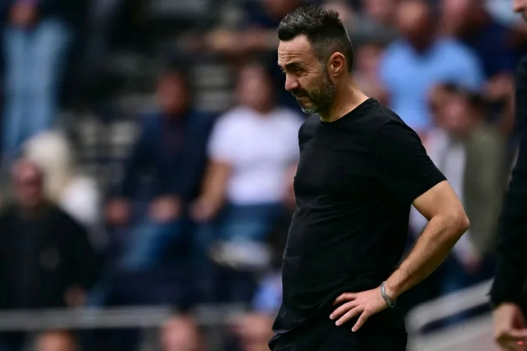 Football manager in black shirt stands with hands on hips beside the pitch