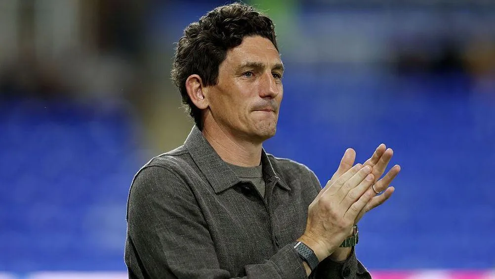 Man in gray shirt clapping with hands raised against a blurred blue stadium background