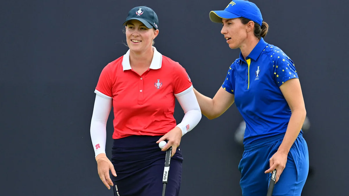 Two female golfers stand together on a course, wearing red and blue team uniforms.