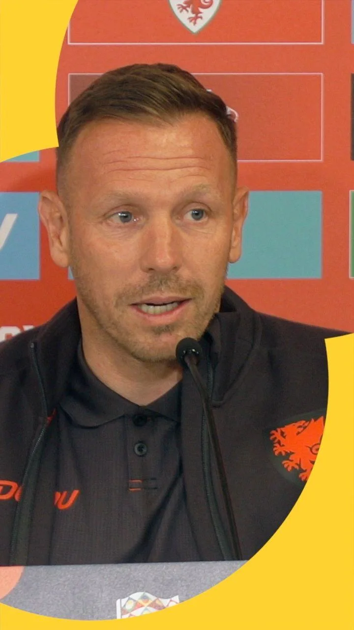 A man in a dark sports jacket speaks at a press conference, seated behind a microphone.