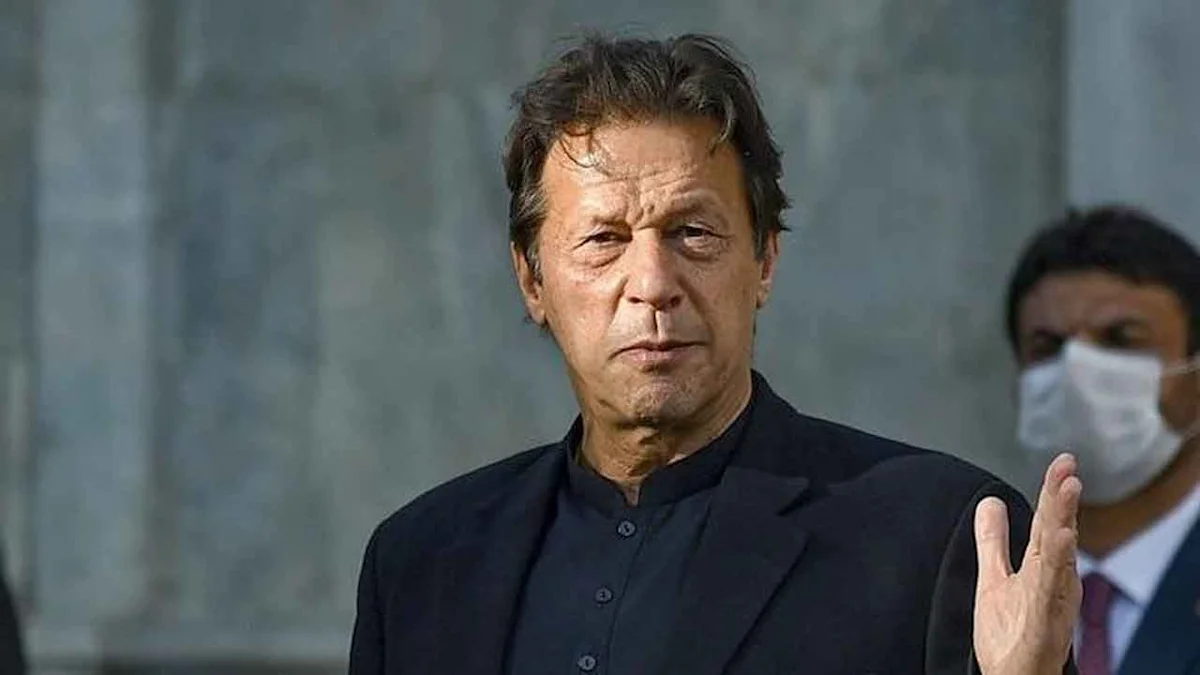 Man in dark traditional clothing raises his hand while standing before a gray wall